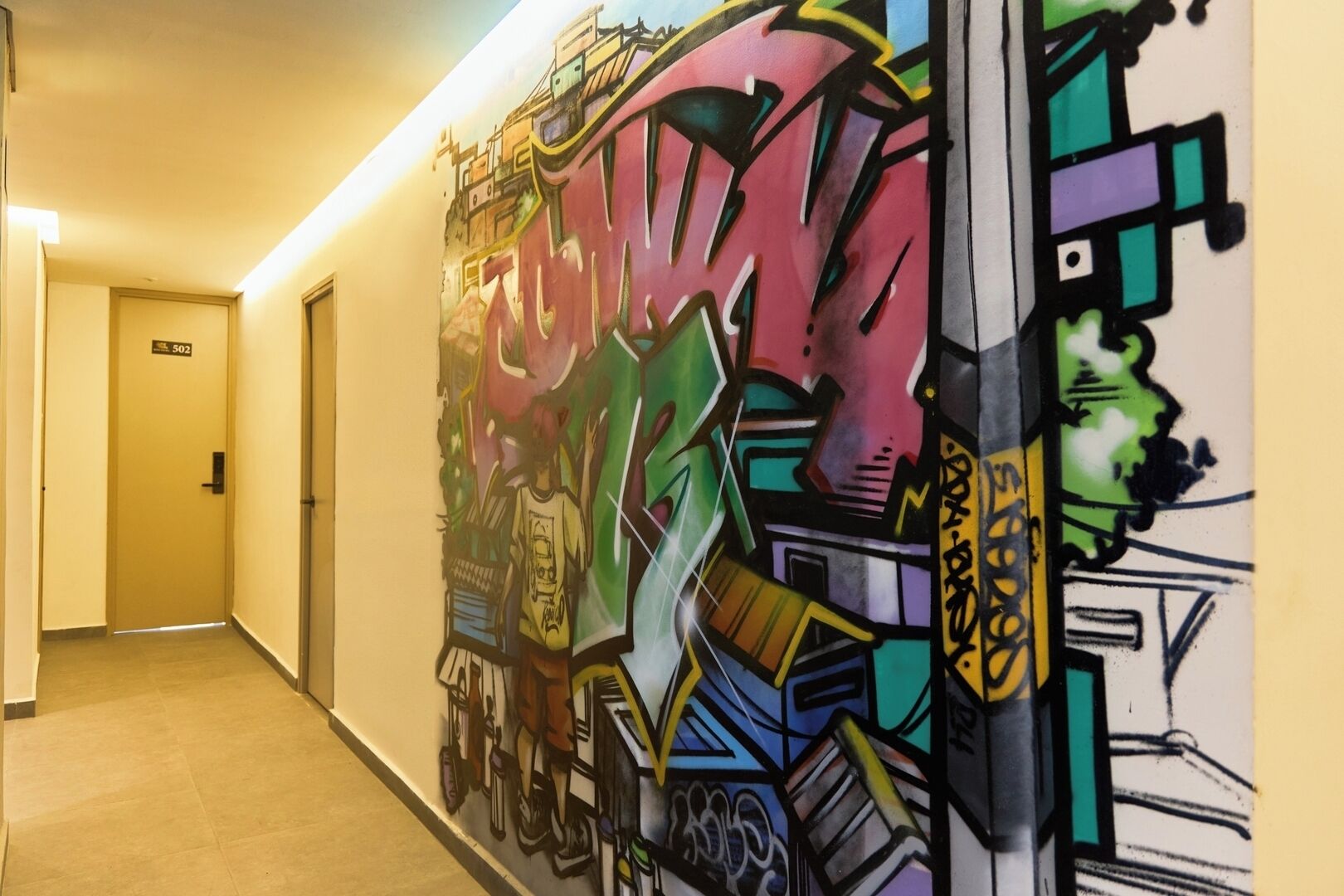 Un pasillo con un mural de grafitis llamativos en la pared, una puerta con el número 502 y un piso de baldosas.