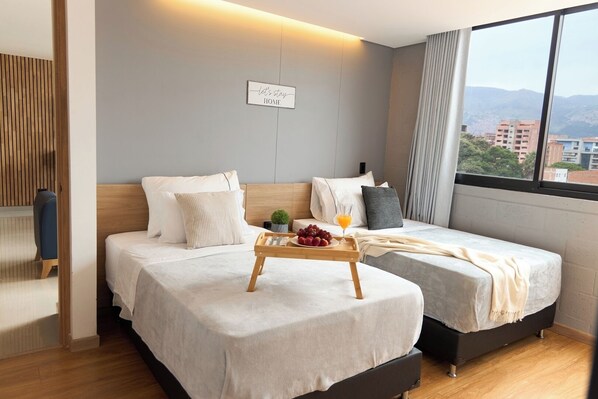 Habitación de hotel con dos camas, cabecero de madera, mesita de noche con una bandeja de fruta y jugo, y vista de la ciudad a través de amplios ventanales.