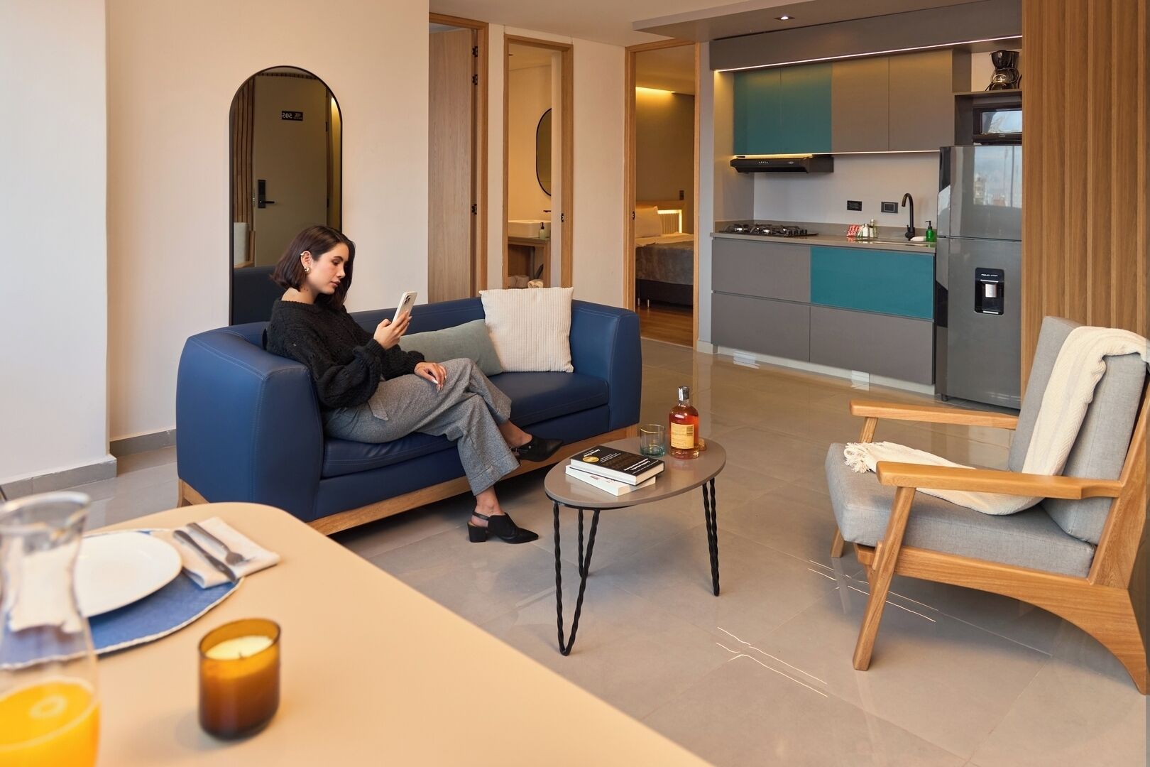 Una sala de estar moderna con un sofá, una mesa de centro y una silla. Una persona está sentada en el sofá usando un teléfono inteligente.