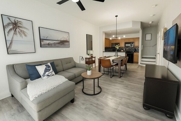 A modern living room with a grey sofa, a small round table, and a TV on the wall. - 3BR Townhome w/ Pool & BBQ Near Disney (Kissimmee)