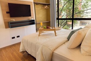 Camera d'albergo con un letto, una TV a parete, un piccolo ripiano con un oggetto decorativo, un vassoio con frutta e bevande, e una finestra vista alberi ed edifici.