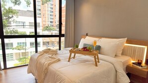 Habitación de hotel con una cama grande, una mesita de noche con lámpara y vistas a edificios a través de amplias ventanas.