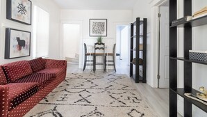 Un pasillo con un sofá rojo, una mesa de comedor con sillas, una estantería negra y obras de arte enmarcadas en las paredes.