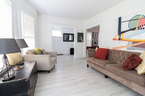 Una sala moderna con un sofá de color claro, un sillón más pequeño y una mesa de centro con una lámpara. Hay un cuadro abstracto grande en la pared y una puerta que da a otra habitación.