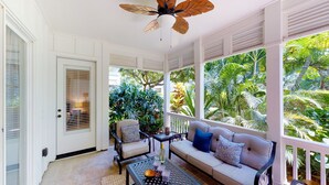 Una sala de estar con ventanales que dan a follaje tropical, con ventilador de techo, sofá con cojines y una mesita de centro.