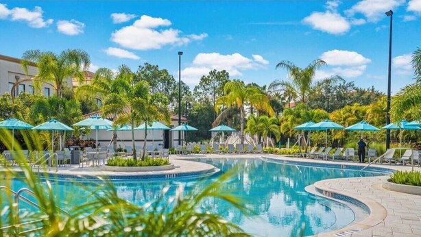 Ein Swimmingpool mit Liegestühlen und Sonnenschirmen, umgeben von Palmen und blauem Himmel.