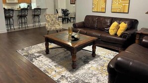 Ein Wohnzimmer mit einem braunen Ledersofa, einem Glastisch und einem gemusterten Teppich. An den Wänden hängen zwei abstrakte Gemälde.