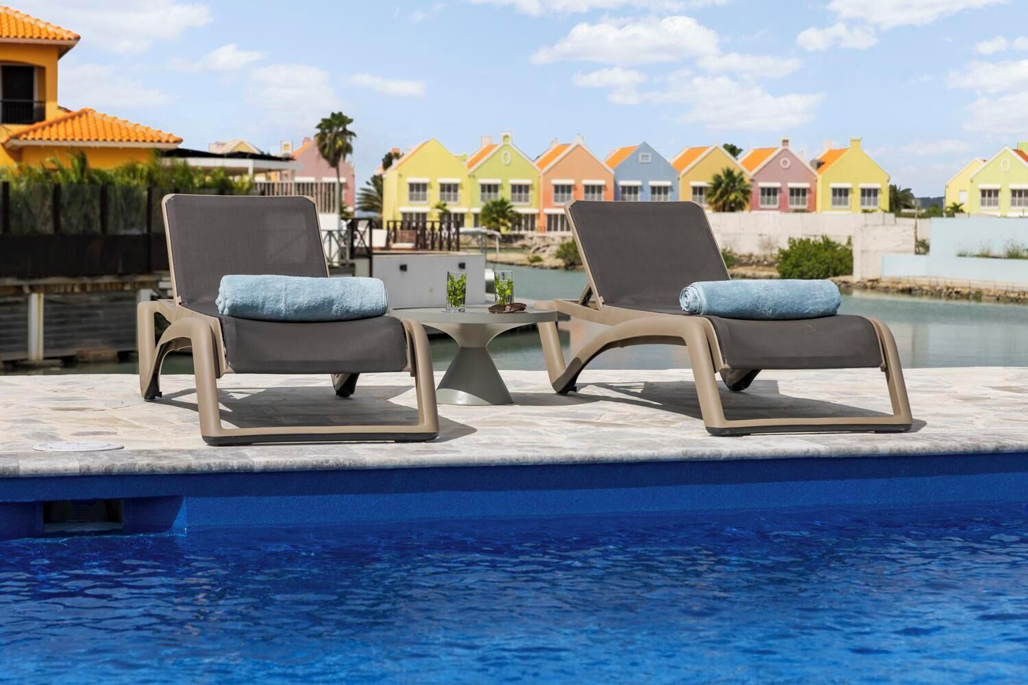 Two lounge chairs with cushions by a poolside, with colorful houses in the background.