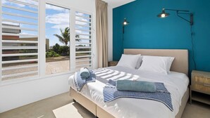 Une chambre avec un grand lit, deux lampes de chevet, une vue par une fenêtre avec stores et un mur d’accent bleu.