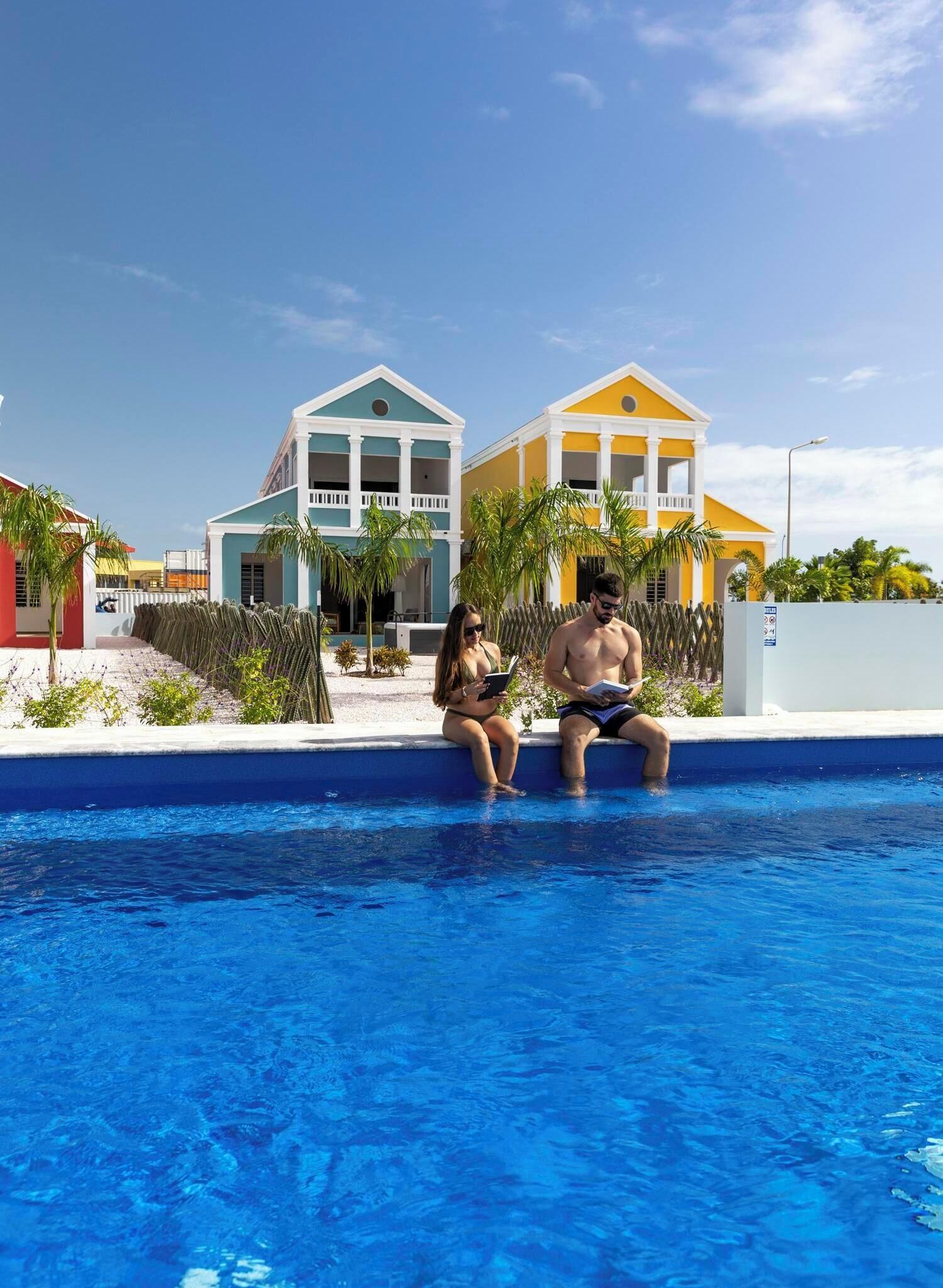 A swimming pool with two people sitting on the edge, a building with a white and yellow facade, and palm trees in the background.