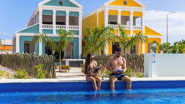 Une piscine avec deux personnes assises au bord, un bâtiment à la façade bleue et jaune, et un jardin de cactus en arrière-plan.