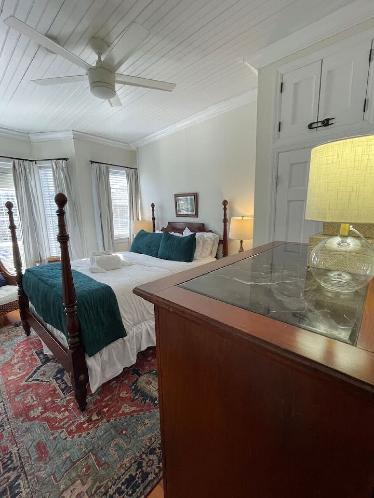 A bedroom with a four-poster bed, a wooden nightstand, a glass-top desk, and a patterned rug.