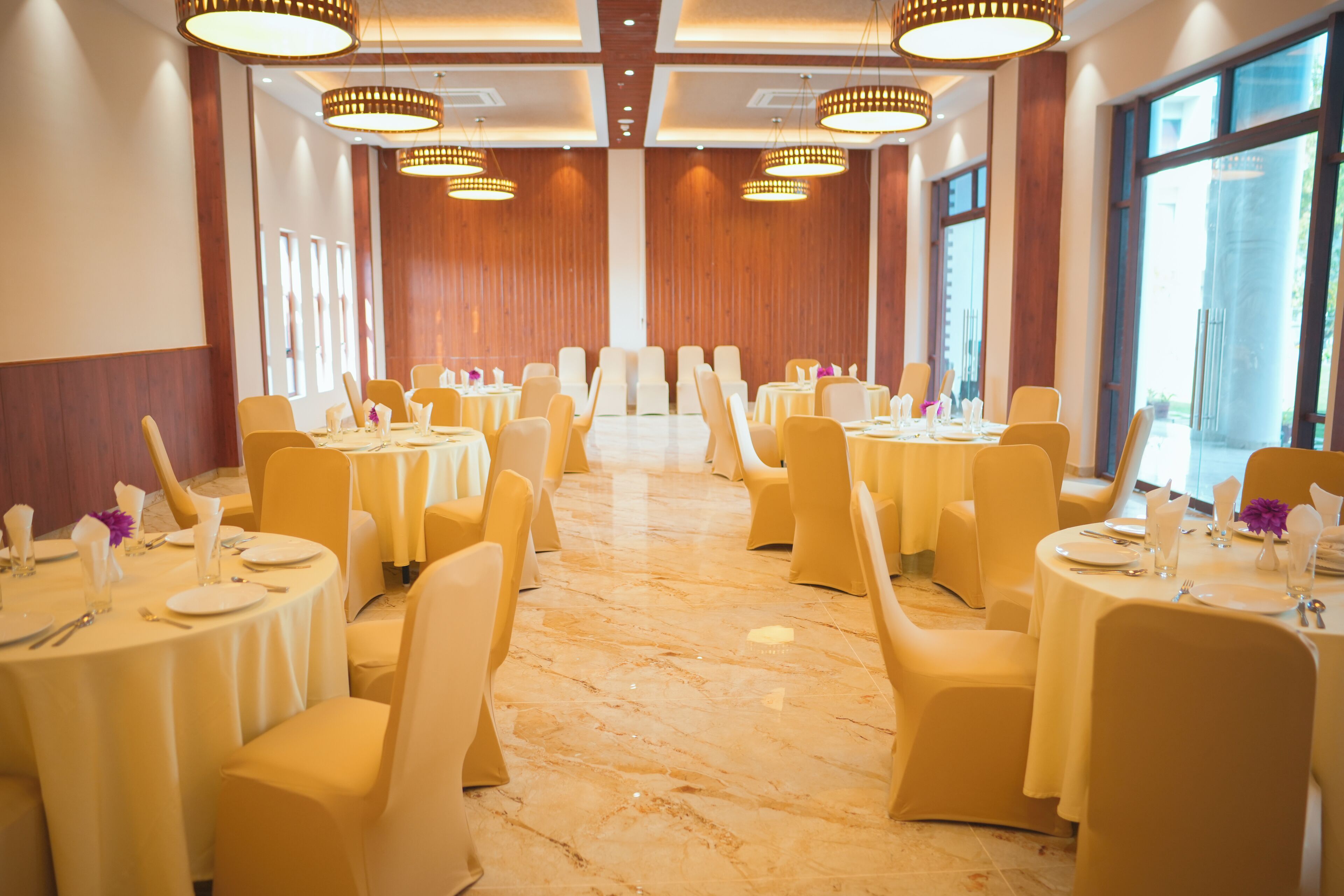 A banquet hall with round tables and chairs, large windows, and chandeliers.
