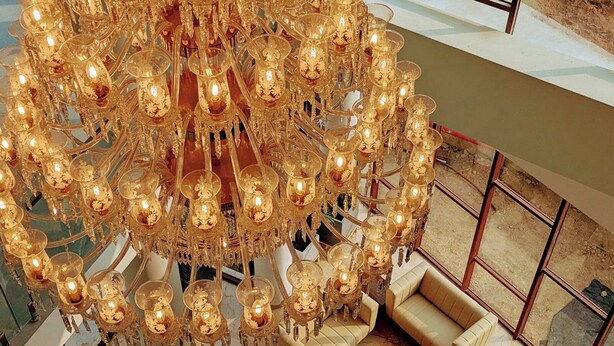 A chandelier with multiple lights, a seating area with sofas and round tables, and a view of the outdoors through large windows. - The Pearl Club Resort (Bolpur)