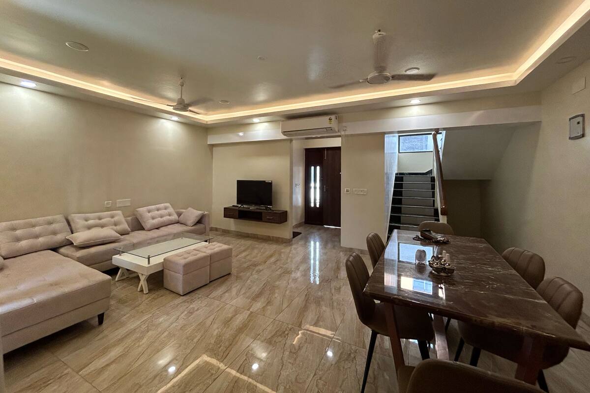 Una sala de estar moderna con sofá, reposapiés, televisor y un comedor con mesa y sillas.