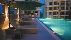 A swimming pool area with lounge chairs and umbrellas, surrounded by a modern building with balconies. - The Pearl Club Resort (Bolpur)