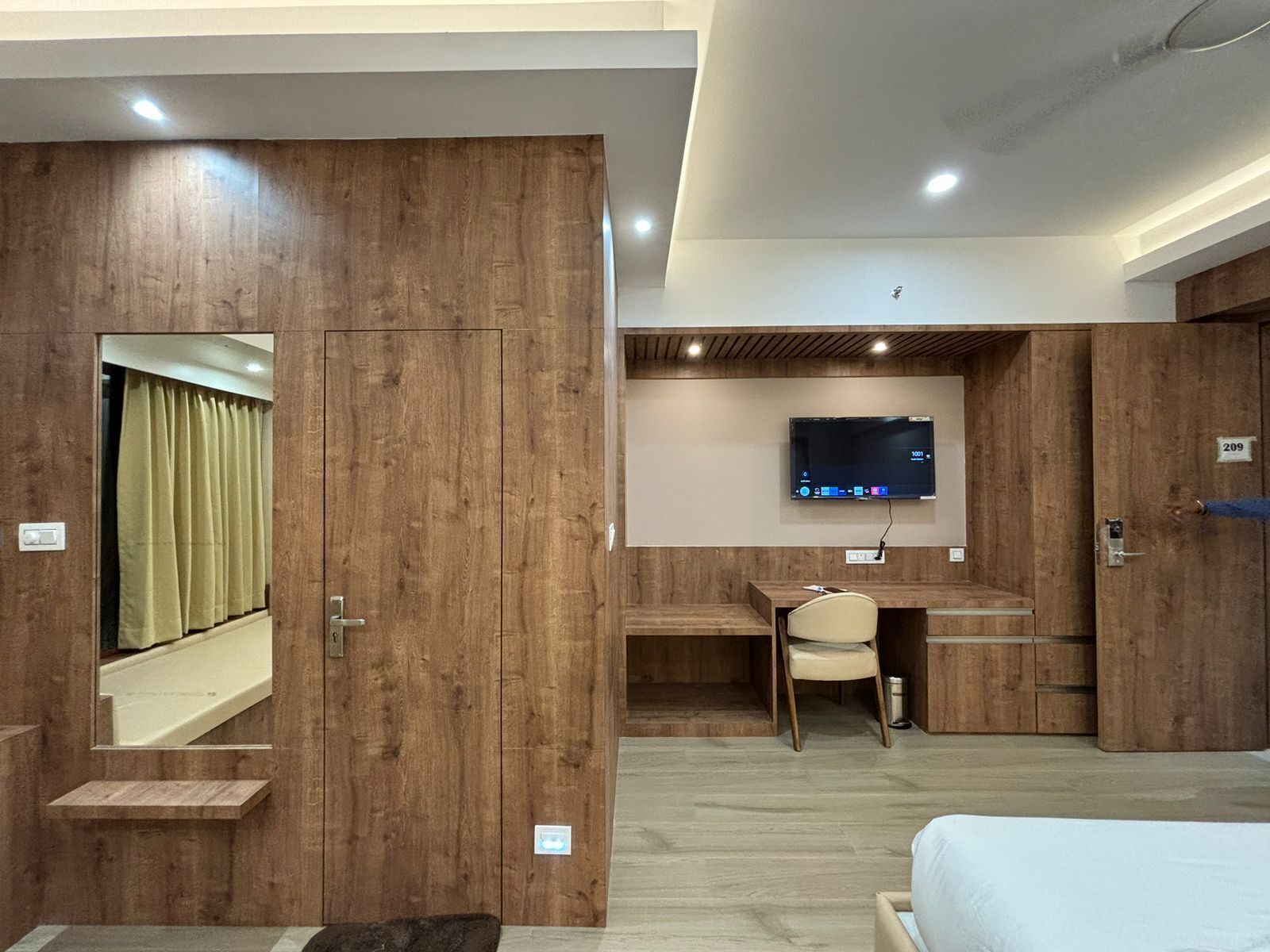 A modern hotel room with wooden walls, a bed, a desk, and a TV mounted on the wall.