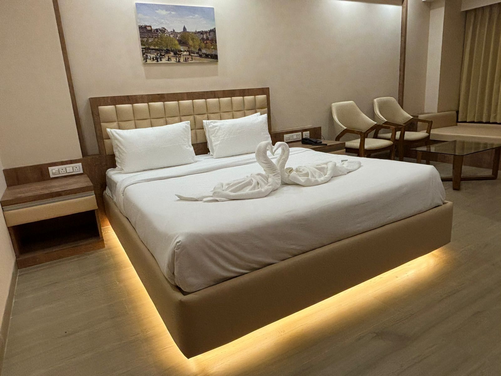 A modern hotel room with a large bed, a nightstand, a desk with chairs, and a painting on the wall.