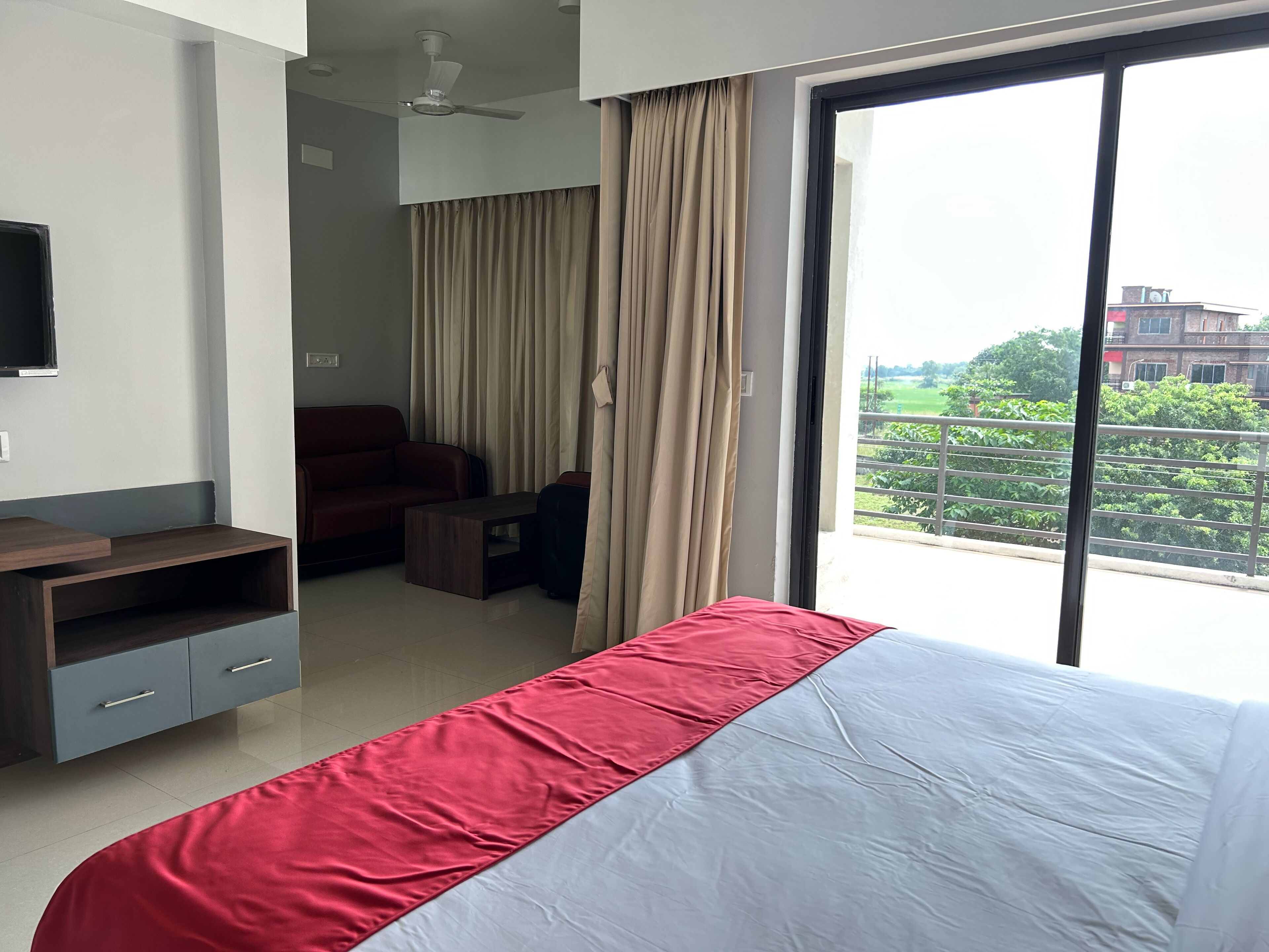 A hotel room with a bed, a desk, a chair, a TV, and a view of greenery outside.