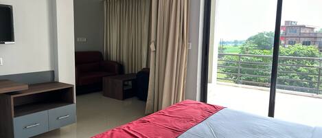 A hotel room with a bed, a desk, a chair, a TV, and a view of greenery outside.