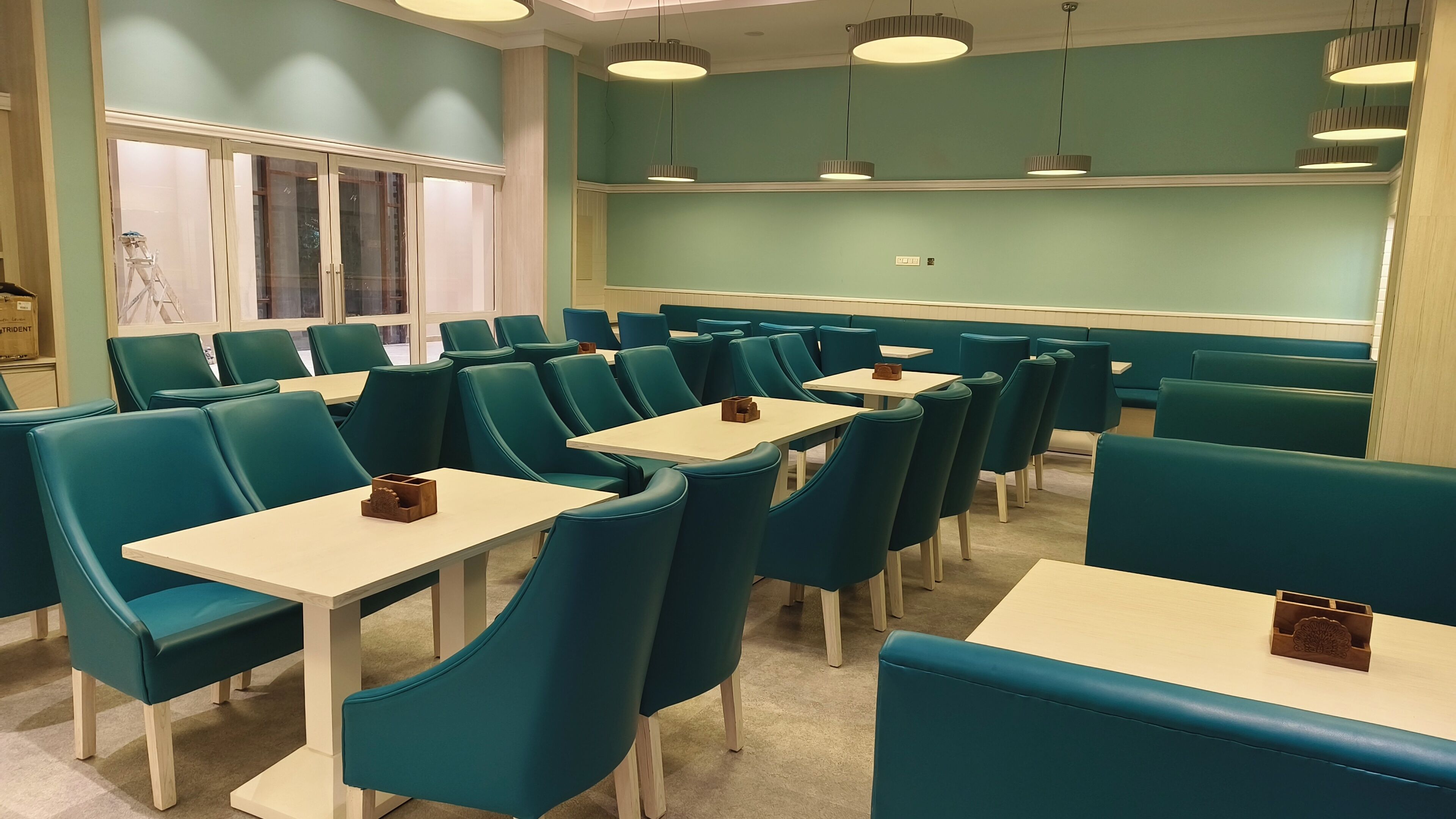 Un restaurante con sillas color turquesa y mesas blancas, con lámparas colgantes y vista a la calle a través de amplios ventanales.