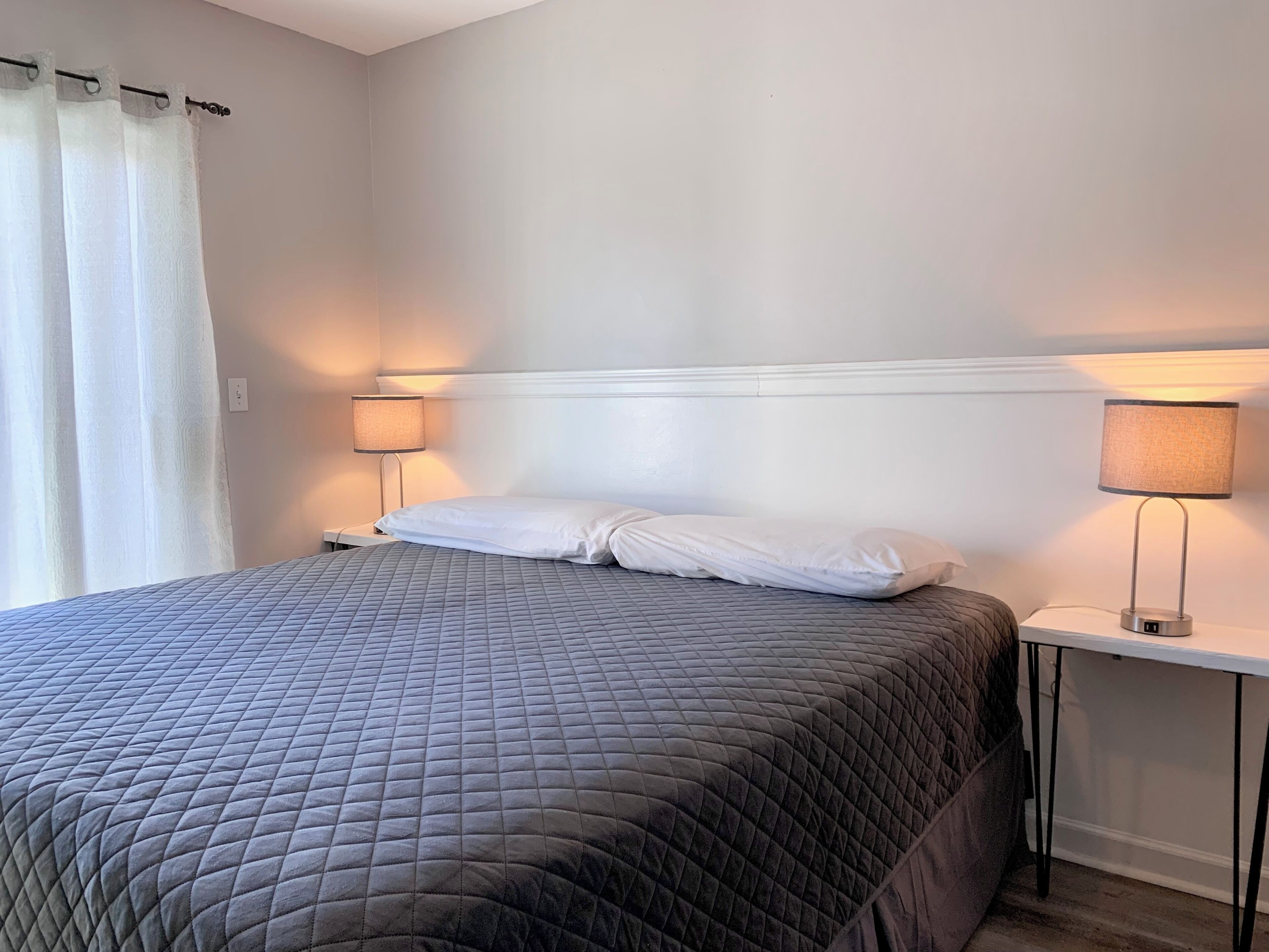 Ein ordentlich bezogenes Bett mit einer gesteppten, grauen Tagesdecke, zwei Nachttischlampen und einem weißen Kopfteil.