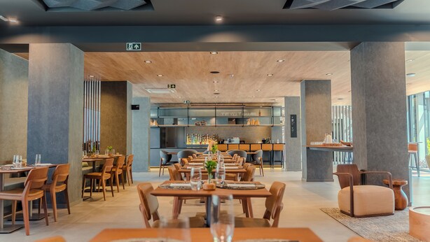 A modern dining area with wooden tables and chairs, a bar area in the background, and a ceiling with geometric patterns. - Laghetto Stilo Garden (Gramado)