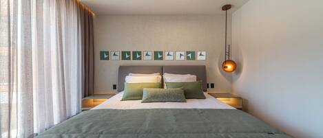 A neatly made bed with green bedding, a headboard with multiple pillows, a bedside table with a lamp, and a window with sheer curtains.