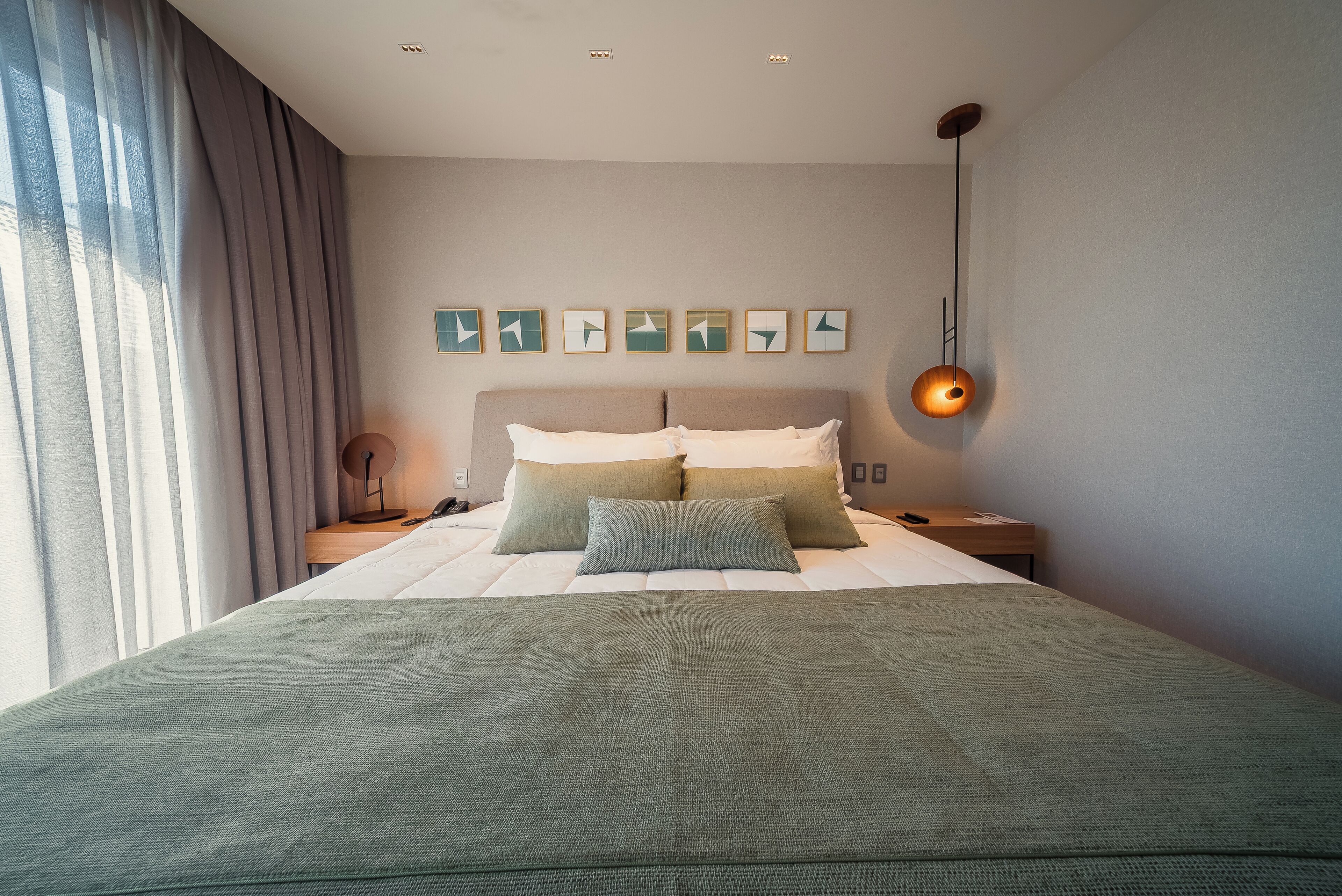Una cama bien hecha con una colcha verde, una mesita de noche con lámpara y una pared decorada con arte abstracto.