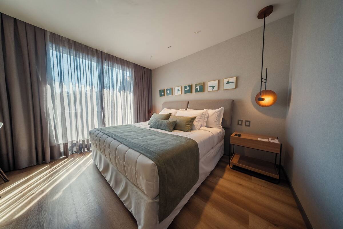 A modern bedroom with a large bed, a bedside table, a hanging light fixture, and a window with curtains.