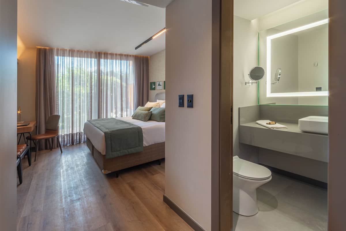 A modern hotel room with a wooden floor, a bed with green bedding, a desk with a chair, a large window with curtains, and a bathroom with a mirror and sink.