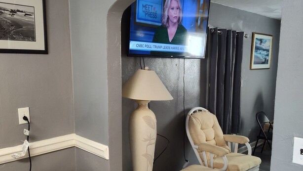 A living room with a television displaying a news channel, a beige armchair, a floor lamp, and a trash can. - Cozy 5-bdrm hm, Loaded Kitchen, w/ Patio 
Deck & Grill, WiFi in pleasant Norfolk (Norfolk)