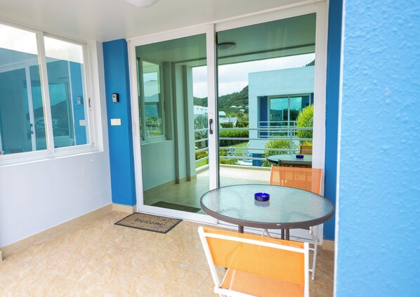 Un balcón con puerta de cristal que da a una terraza, una mesa redonda de cristal con un cuenco azul, y un felpudo.