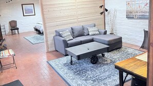 Ein modernes Wohnzimmer mit einer grauen Eckcouch, einem Couchtisch und einem Esstisch. Auf dem Boden liegt ein gemustertes Teppich, und die Decke ziert sich mit Holzbalken.