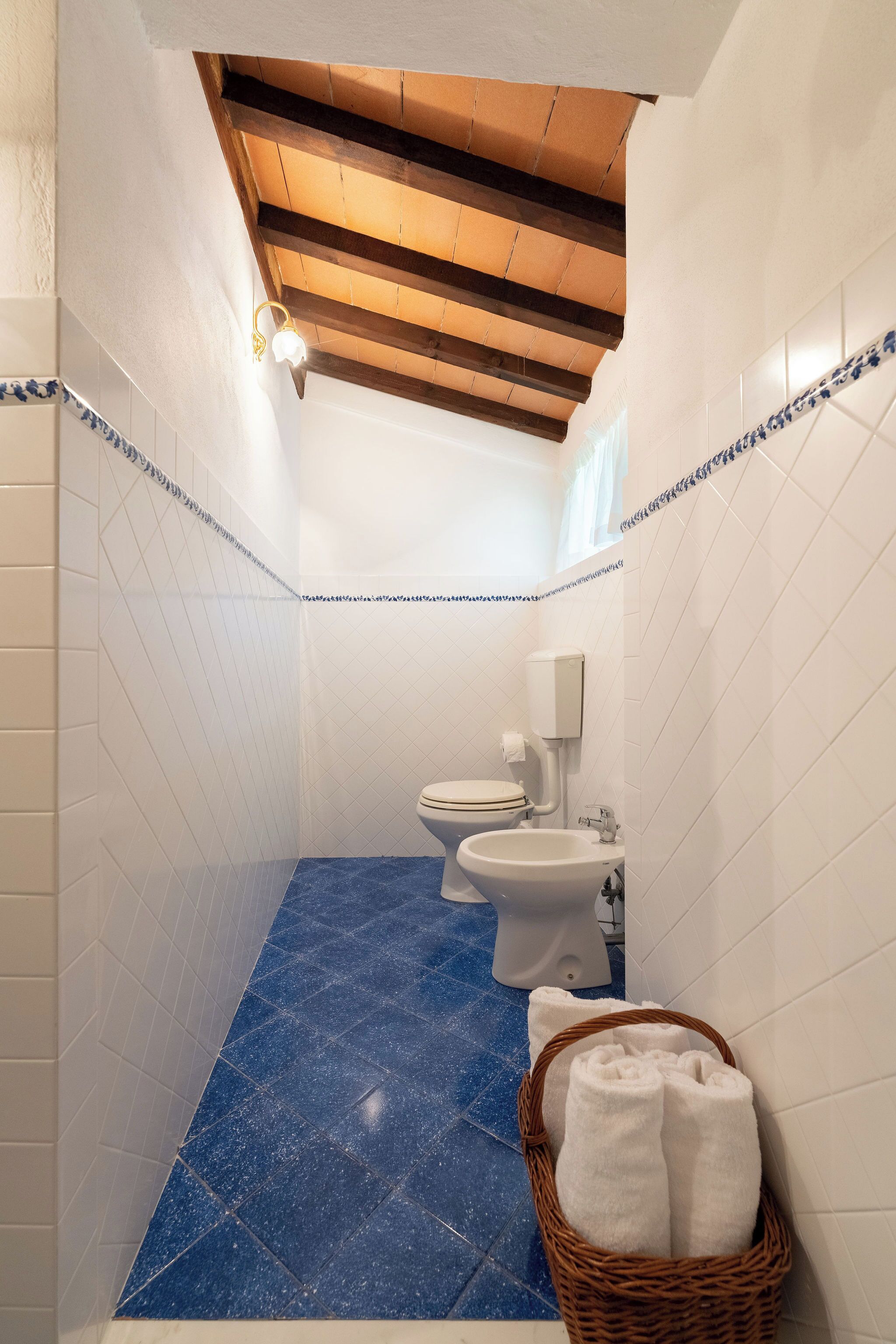 Een kleine badkamer met een blauwe betegelde vloer, witte wanden en een houten balk in het plafond. Er is een wit toilet, een bidet en een mand met witte handdoeken.