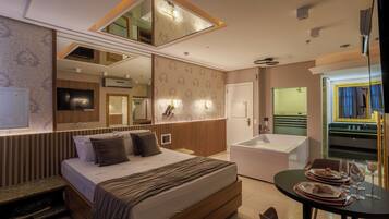 A modern hotel room with a large bed, a separate bathroom with a bathtub, a dining area with a table set for two, and a large mirror above the bed.