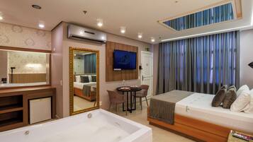 Habitación de hotel moderna con una bañera grande, televisión, una pequeña mesa de comedor y una cama con almohadas.
