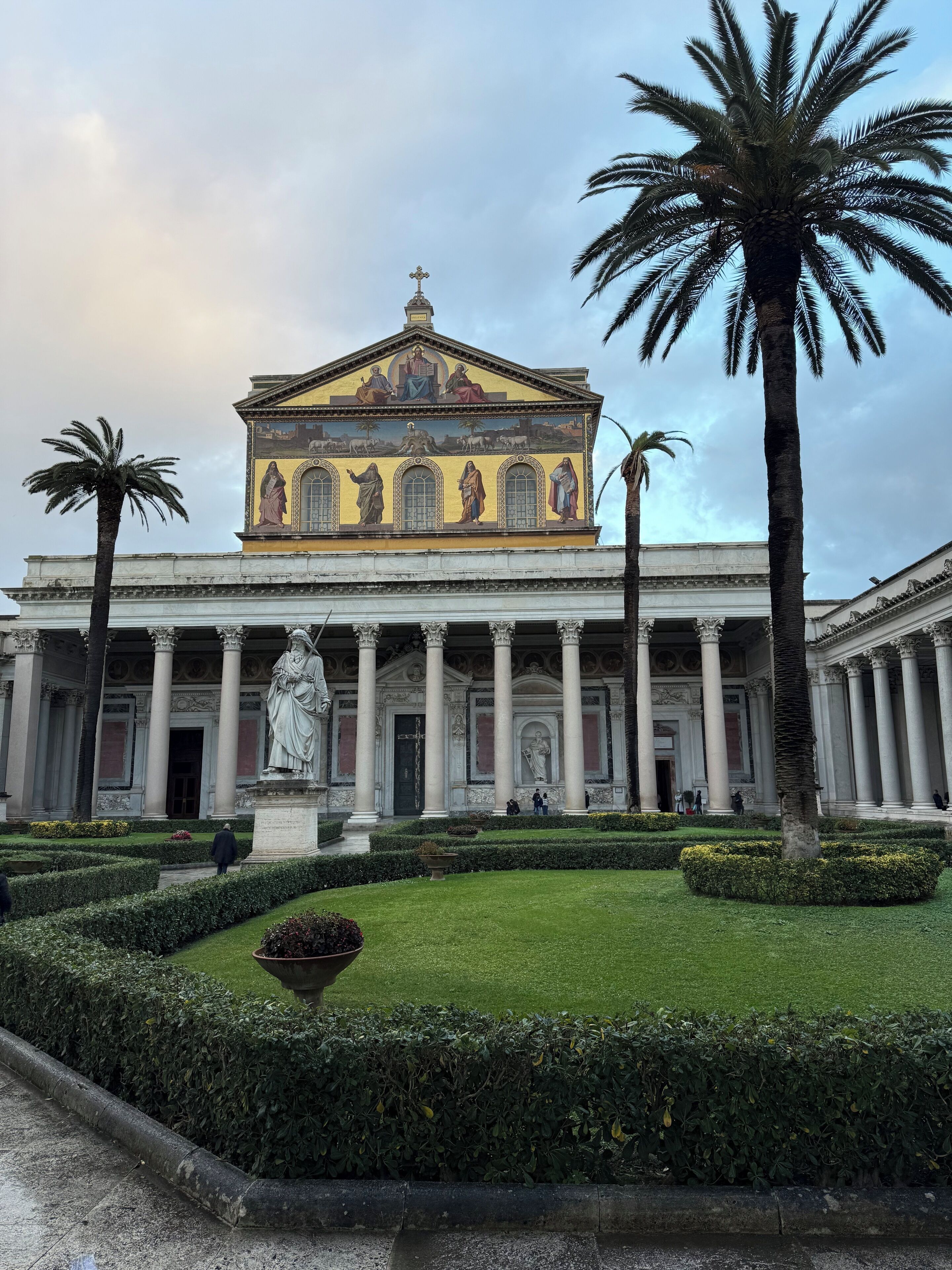 Un edificio clásico con una estatua central, rodeado de jardines cuidados y palmeras.