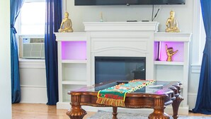 Een woonkamer met een flatscreen televisie boven een open haard, een houten salontafel met een kleurrijke plaid, en blauwe gordijnen.
