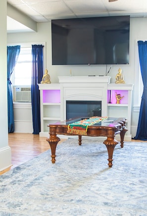 A living room with a flat-screen TV mounted above a fireplace, a wooden coffee table with a colorful throw, and blue curtains. - Fall River Stay Stylish 2BR • Free Parking (Fall River)