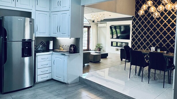 Una cocina moderna con armarios blancos, un refrigerador de acero inoxidable y un comedor con pared de estampado negro y dorado, y una araña.