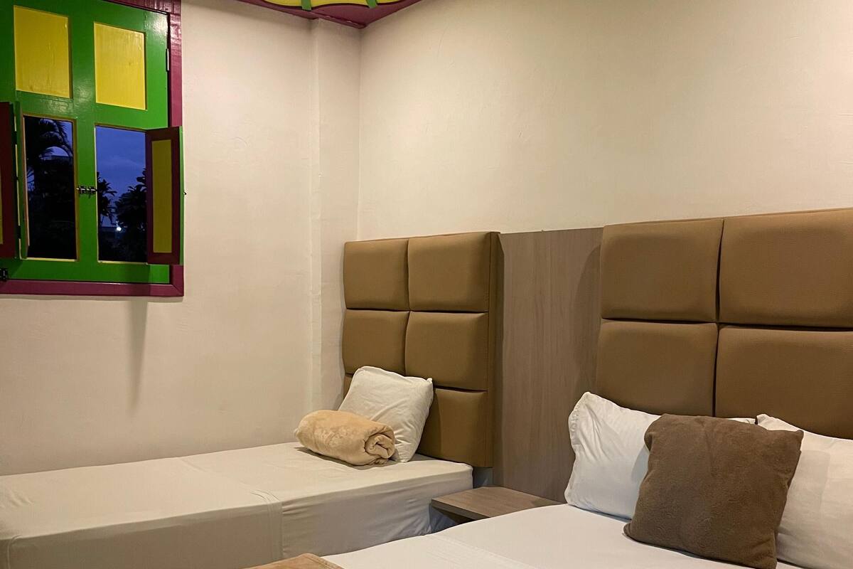 Une chambre d’hôtel avec deux lits, une fenêtre encadrée de vert et un plafond à rayures jaunes.