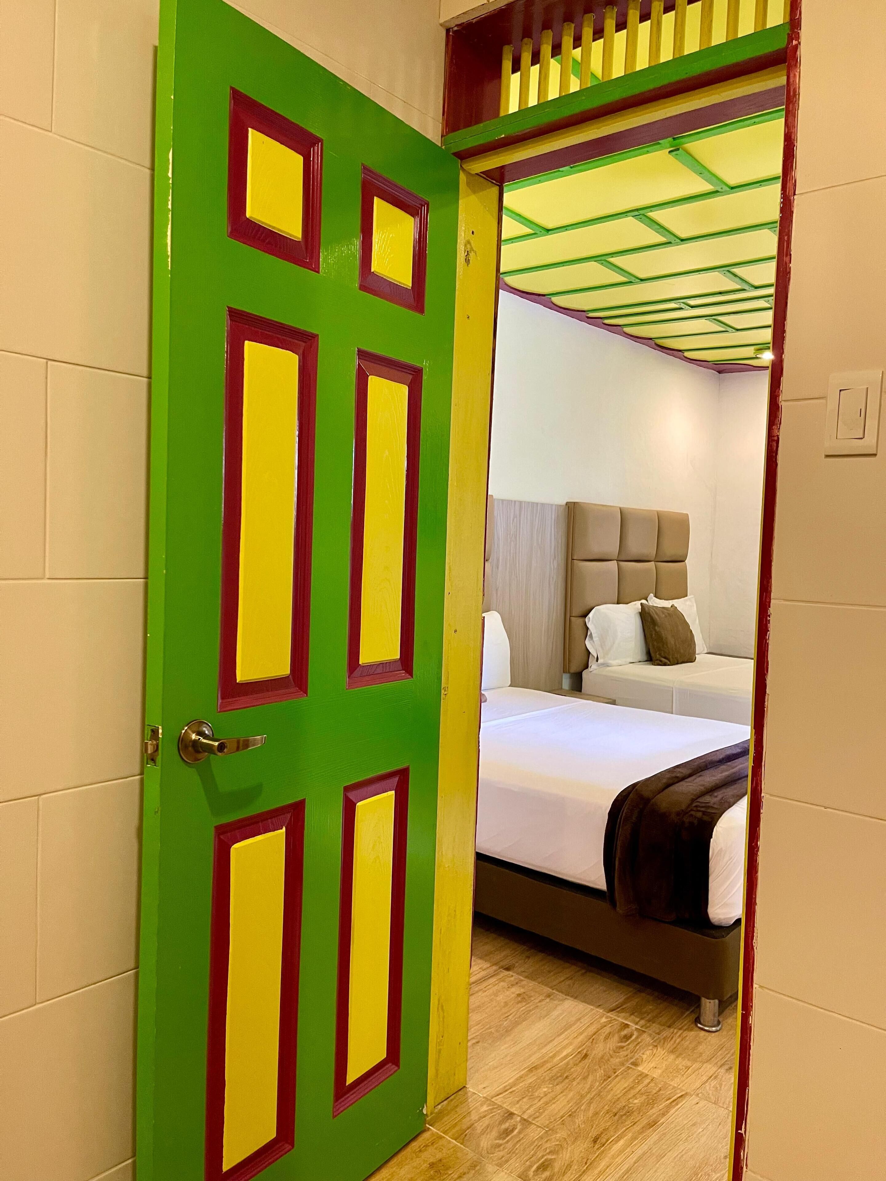 A room with a green door with red and yellow accents, a bed with a headboard, and a wooden floor.