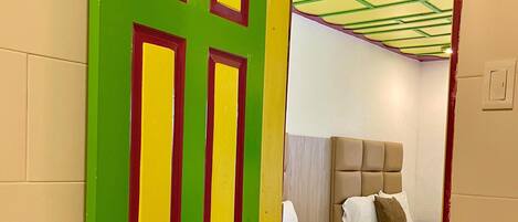 A room with a green door with red and yellow accents, a bed with a headboard, and a wooden floor.