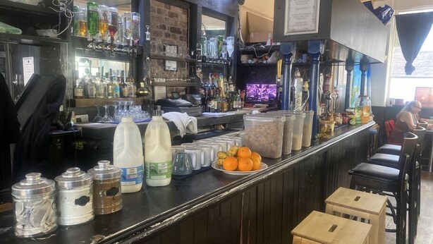 Zona de bar amb un mostrador que mostra diverses ampolles i recipients, una pantalla de televisió i una zona de seients amb taules i cadires.