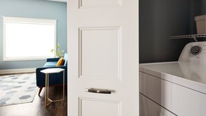 A room with a large window, a blue sofa, a small round table, and a laundry area with a washing machine. - Pickwick Places 201 MSU/Rountree (Springfield)