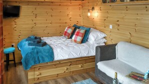 A cozy wooden cabin room with a bed, a sofa, a television, and shelves with various items. - Brockalee Farm Retreat (Bardon Mill, Near Hexham)