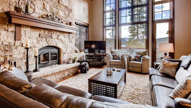 A spacious living room with a stone fireplace, leather sofas, a coffee table, and large windows offering a view of trees. - Breck_snowy_point_chalet (Breckenridge)