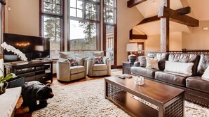 A spacious living room with a large TV, comfortable seating, and a view of trees through large windows. - Breck_snowy_point_chalet (Breckenridge)