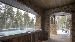 Ein Whirlpool in einer rustikalen Holzhütte mit Steinwänden und Holzbalken mit Blick auf einen verschneiten Wald.